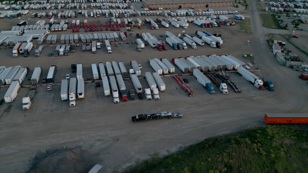 Aerial wide shot of a semi-truck exiting a U.S. truck-stop parking lot