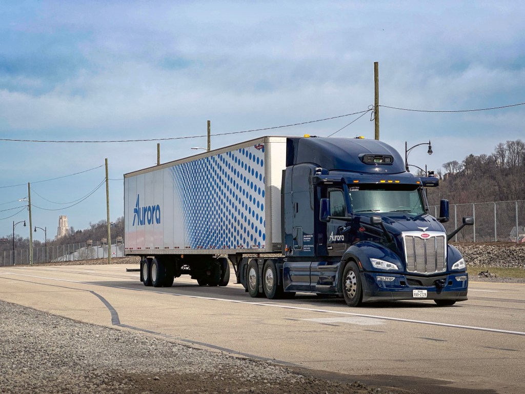 A blue semi-truck with a trailer featuring a dot pattern, driving down a wide road with utility poles in the background.